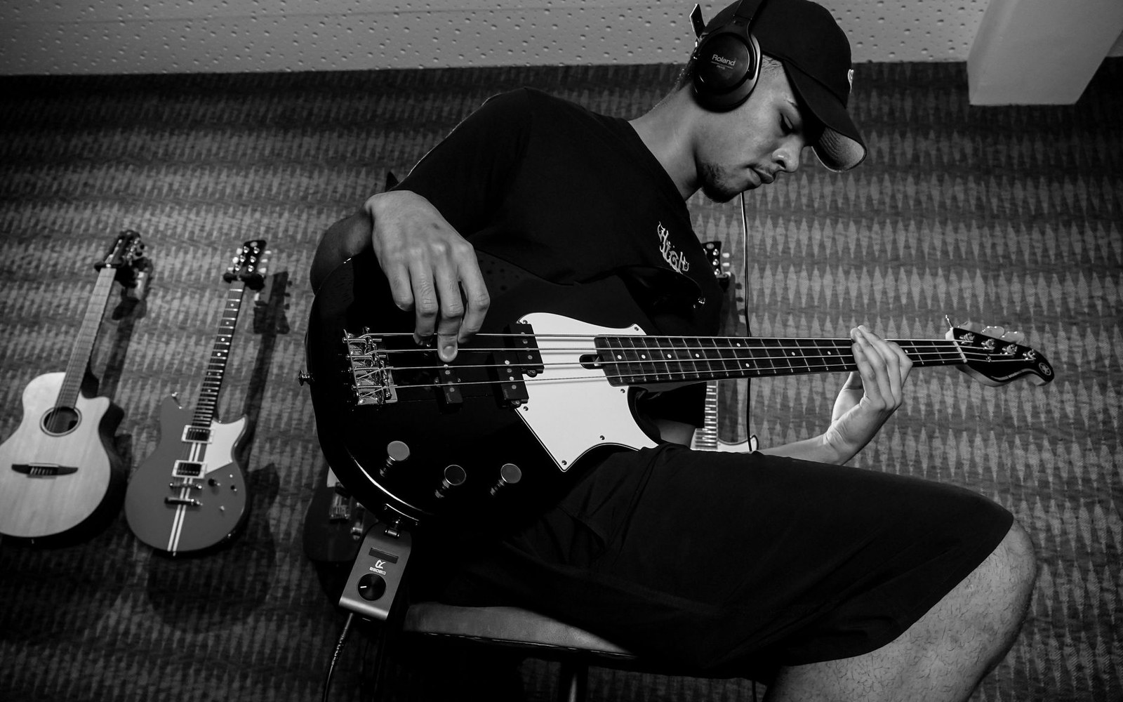 Man wearing headphones plays a bass guitar while seated near two other guitars