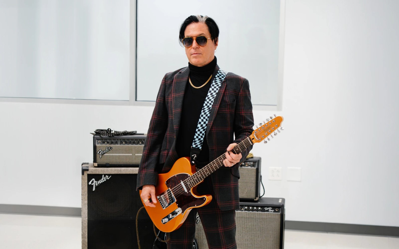 Man in plaid suit and sunglasses holding an orange electric guitar beside Fender amplifiers