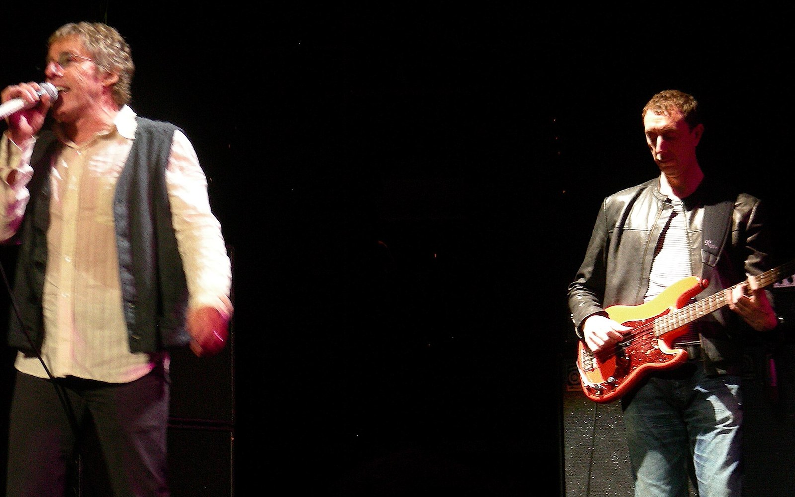 Singer holding a microphone performs beside a bassist playing an electric bass guitar