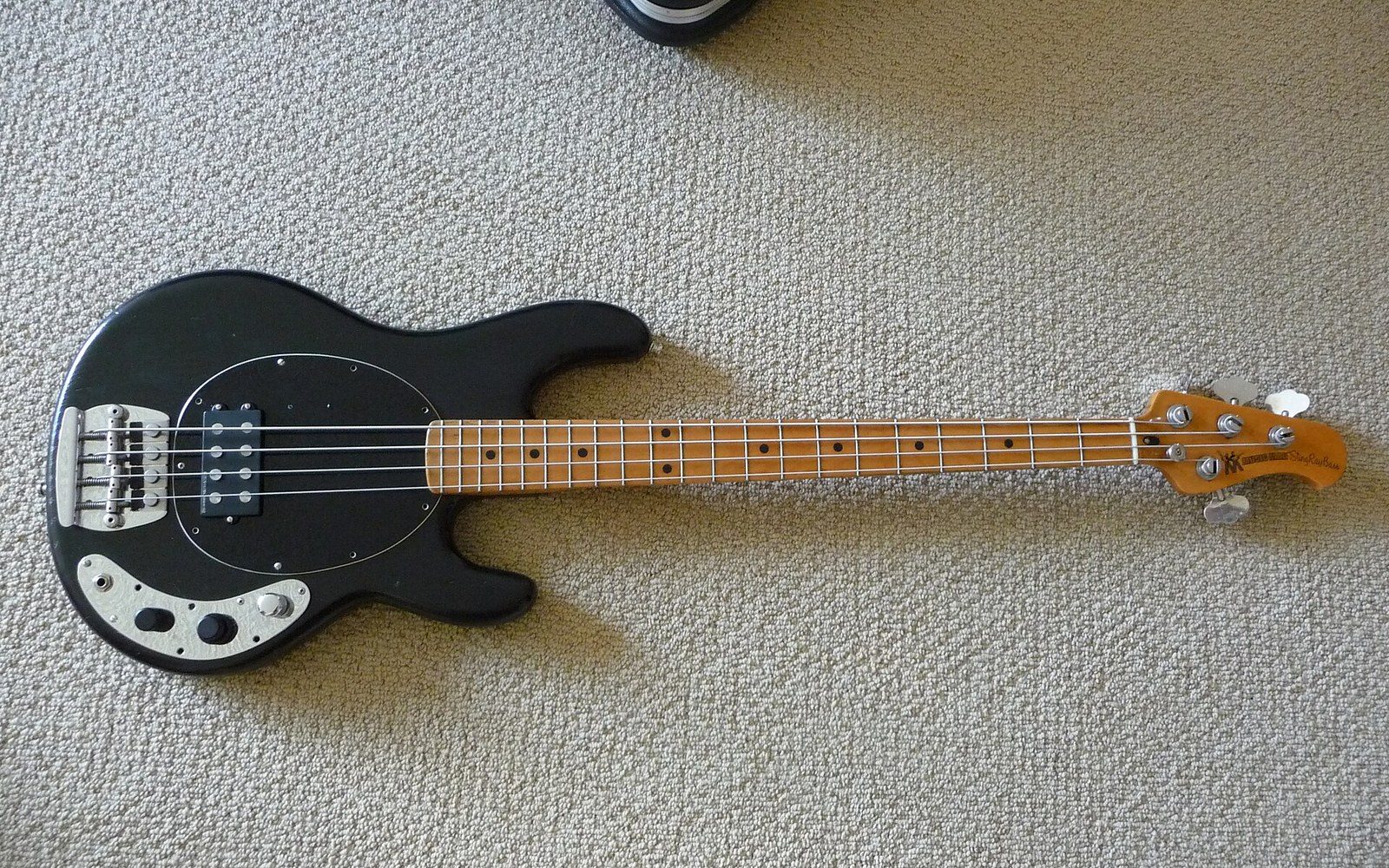A black electric bass guitar with a maple neck lies on a carpeted floor