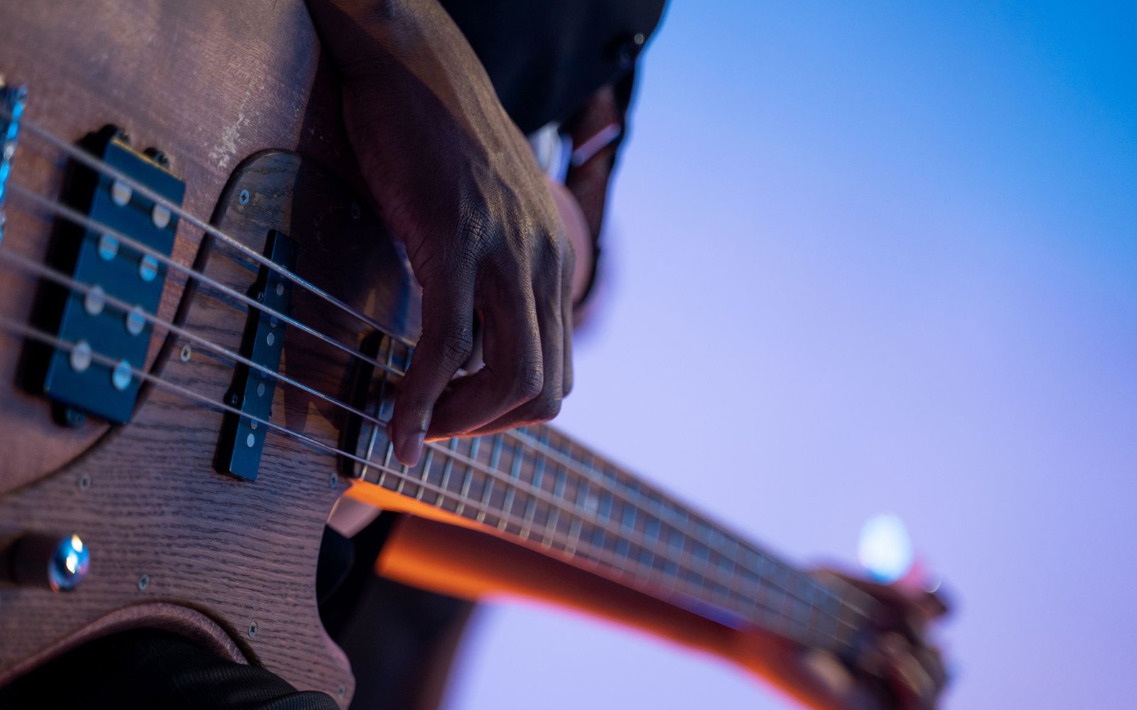 An adult player's right hand positioned over the split-coil pickup of a sunburst Precision Bass, fingers at rest between the pickup and the bridge, stage light from the left