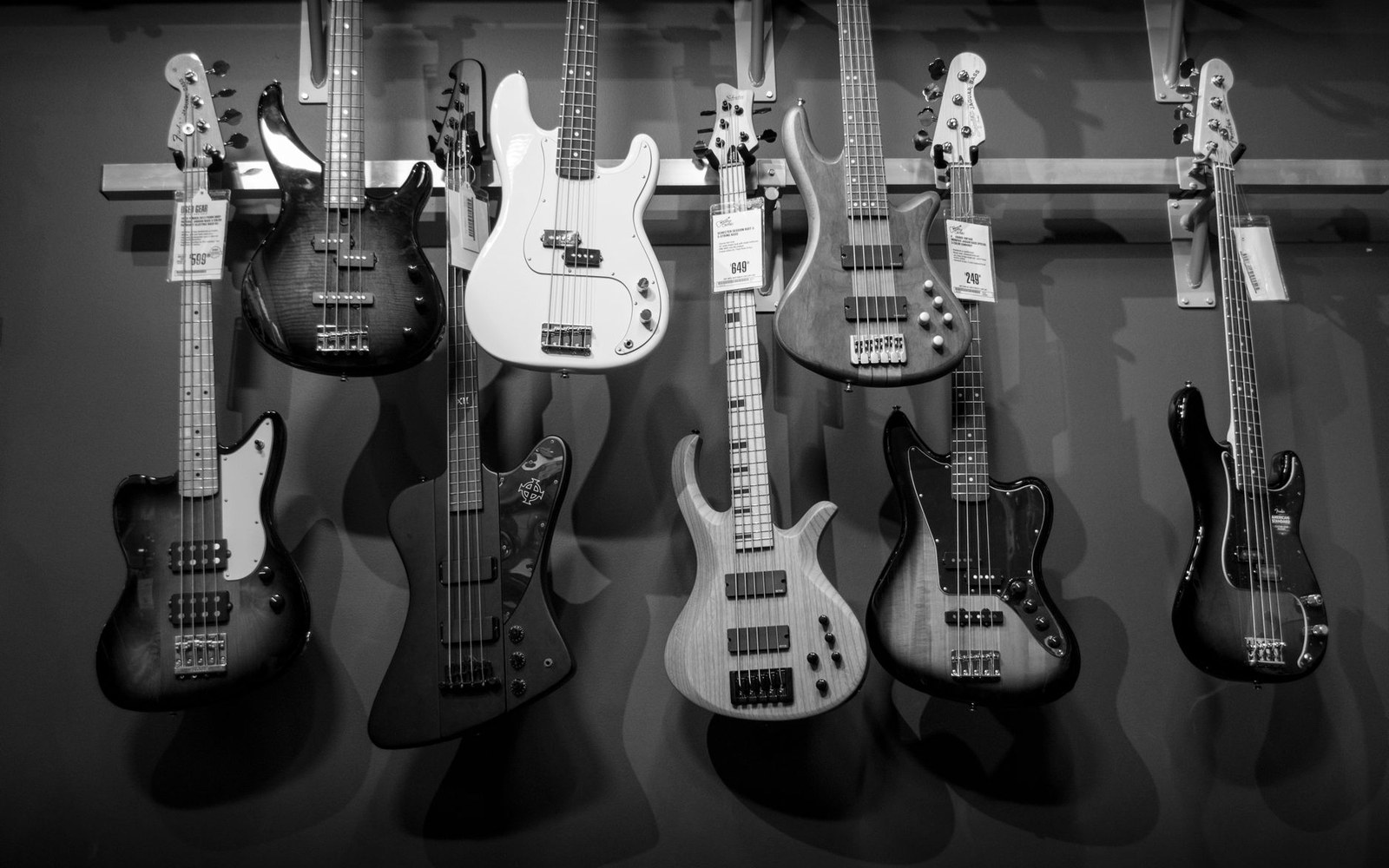 A wall of bass guitars hanging in a music shop — Precision, Jazz, StingRay and Rickenbacker body shapes visible — an adult member of staff in the background